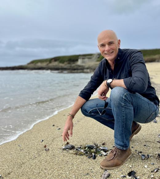 Smiling man crouched on sandy beach near calm shoreline.