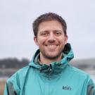 Outdoor portrait photo, smiling man in teal rain jacket with blurred shoreline